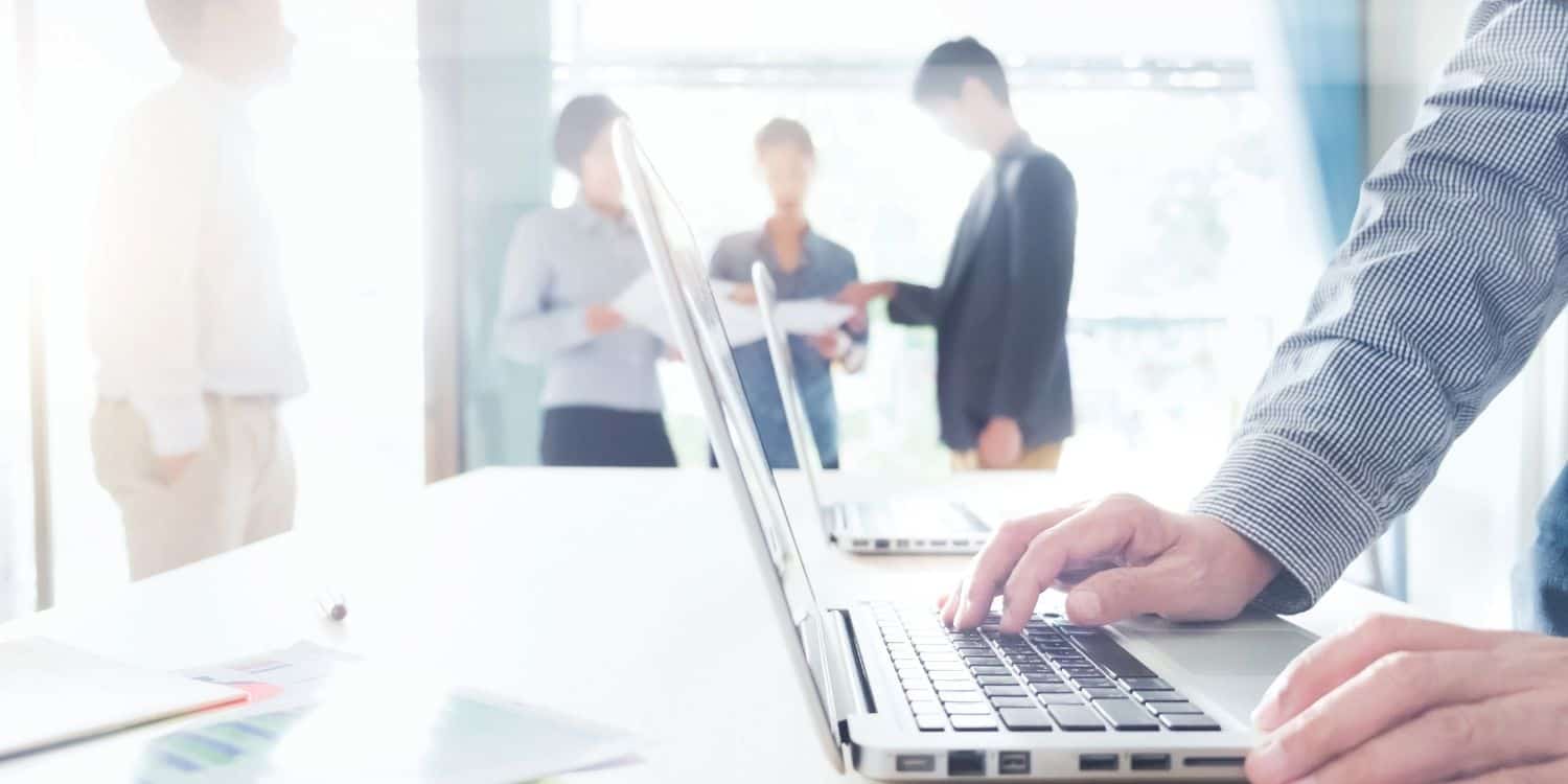 photo of a man's hands on a laptop with a meeting happening further back in the background