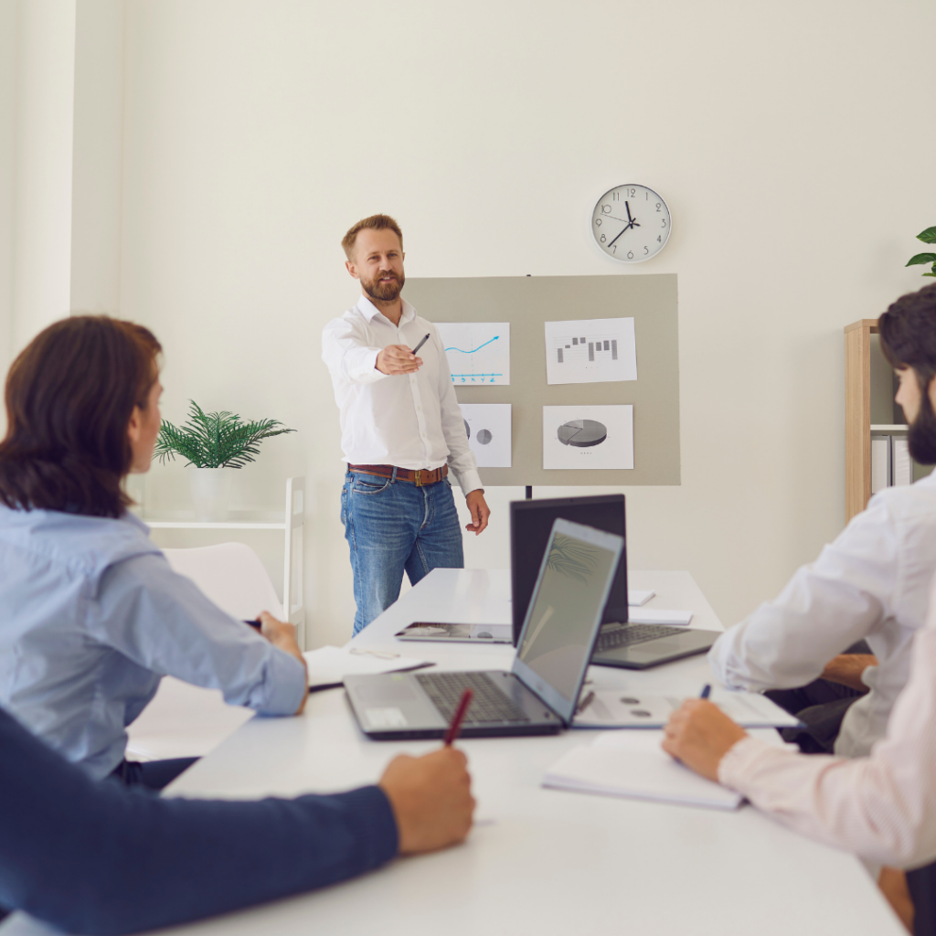 image of white male using hands to gesture to make a point standing in front of a group of people in a meeting